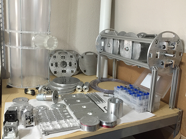 Picture of an assortment of machined parts for the HAWK tool which can be seen in images below.  On the left is a clear 11 inch diamter and 4 ft long chamber with an access port to feed the wires into the chamber.  On the right is one of the assembled scaffoldings of the internal modules of the hawk tool.
